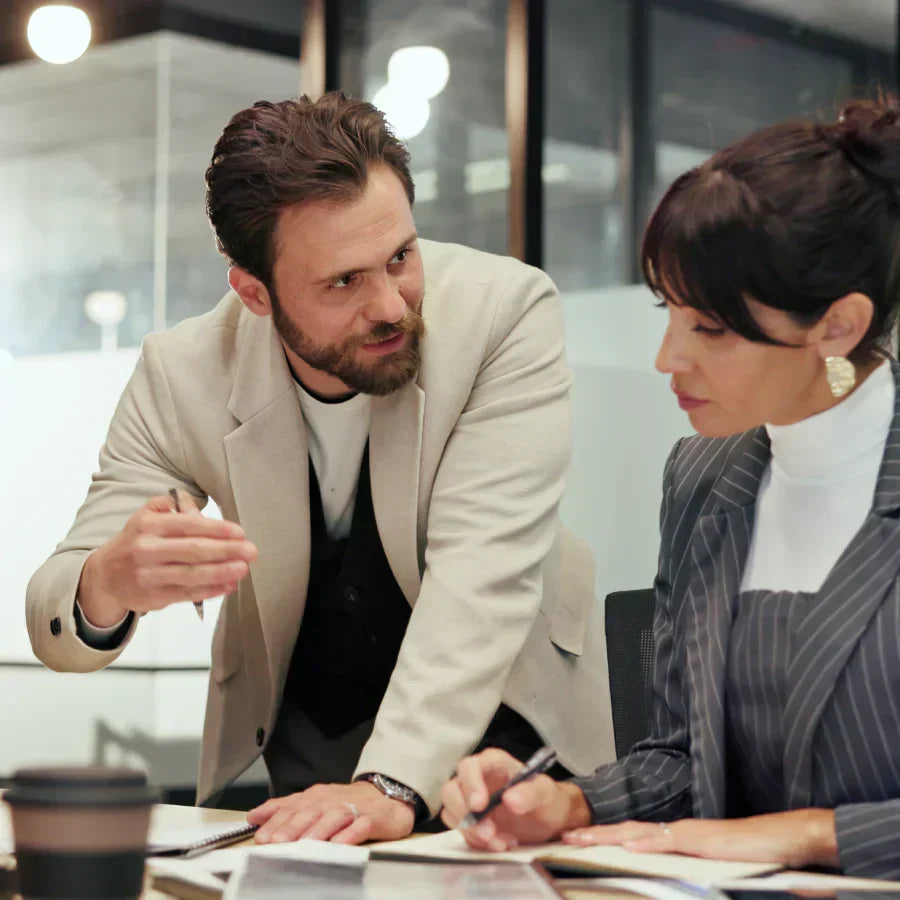 Two professionals collaborating in an office, with a woman wearing a silver Plaud NotePin on her blazer lapel.