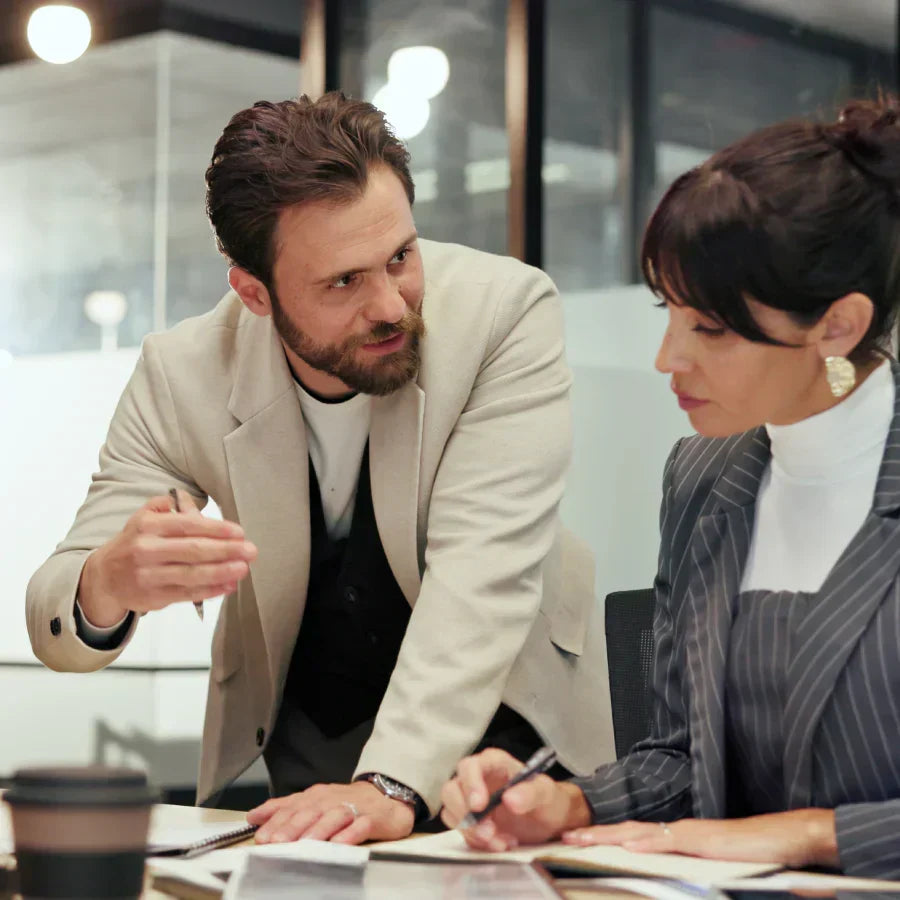 A man in a light jacket gestures while speaking to a woman writing in a notebook during a business meeting.