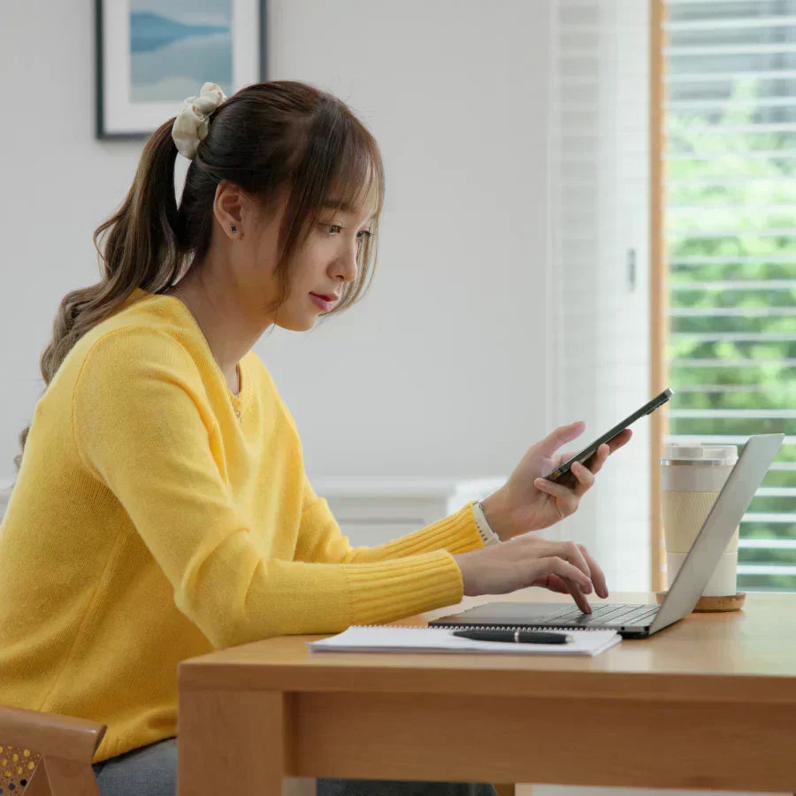 Woman in a yellow sweater using a laptop and holding a smartphone with a black Plaud Note attached to the back.