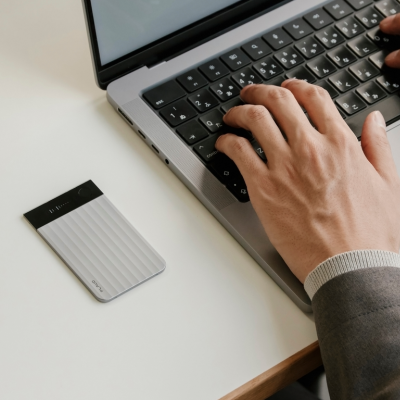 Silver Plaud Note Pro with a ribbed texture and screen sits on a desk next to a person typing on a laptop.
