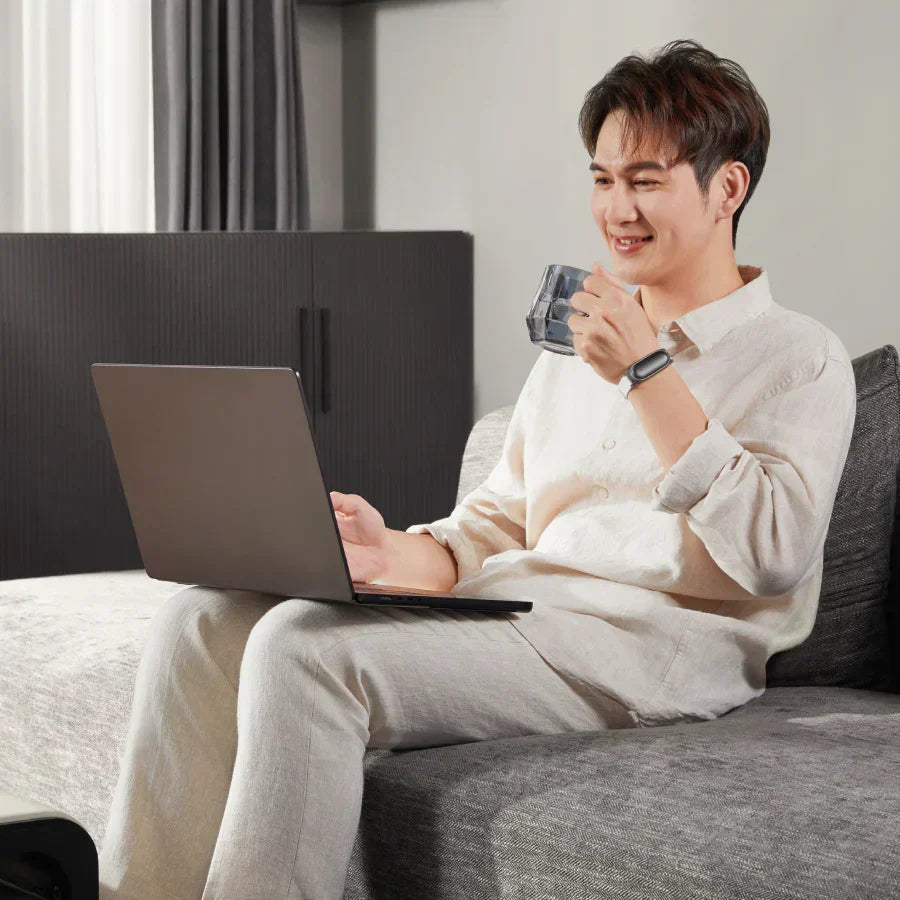 Man on a sofa using a laptop and wearing a black Plaud NotePin on a white wristband.