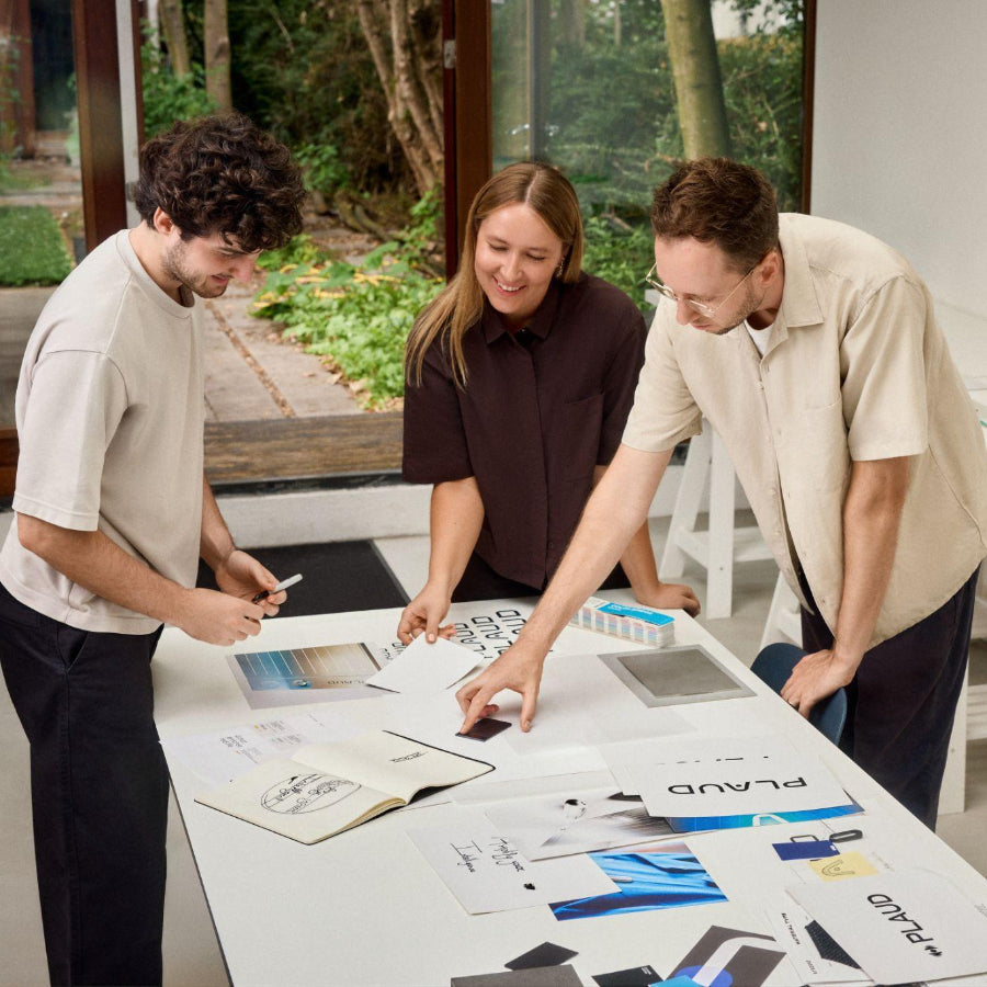 Three people reviewing design sketches and a black Plaud Note on a table in a creative workspace.