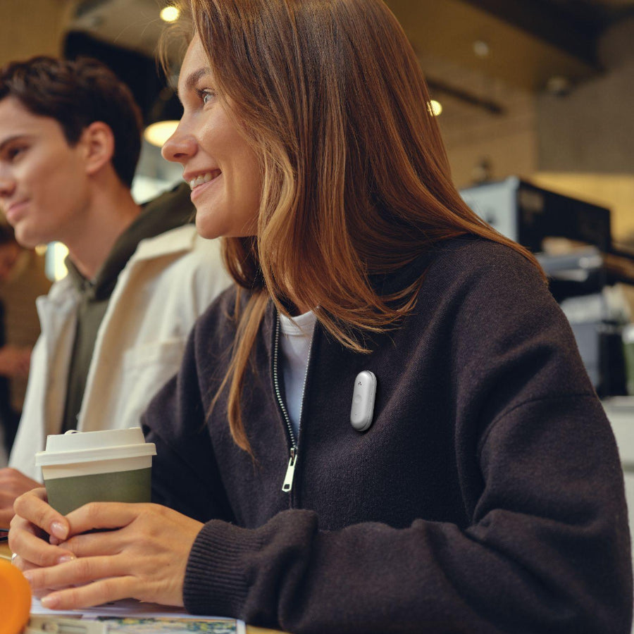 A woman in a cafe wearing a silver Plaud NotePin S on her dark sweater while holding a coffee cup.