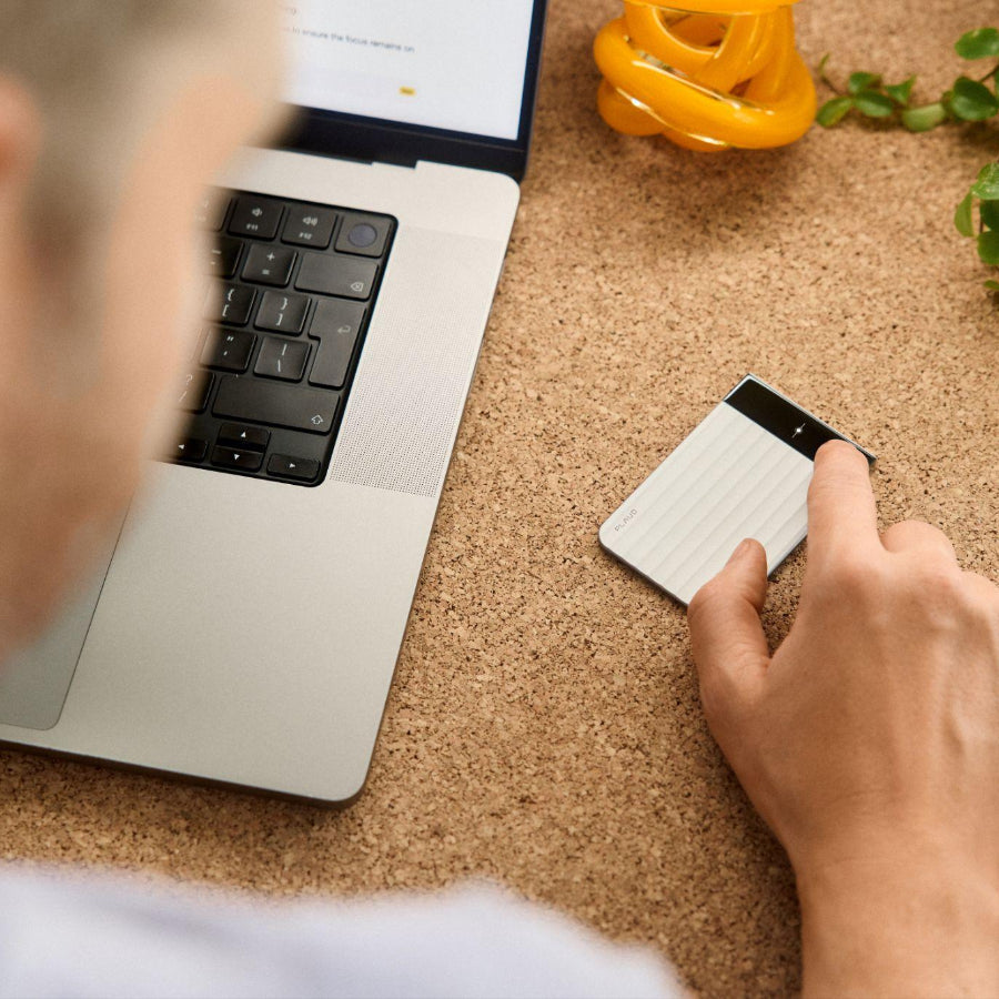 Person using a silver Plaud Note Pro with a display screen on a cork desk next to a laptop.
