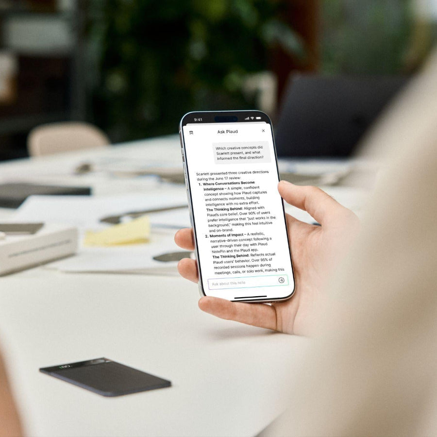 Hand holding a phone with the Plaud App interface next to a black Plaud Note Pro on an office desk.