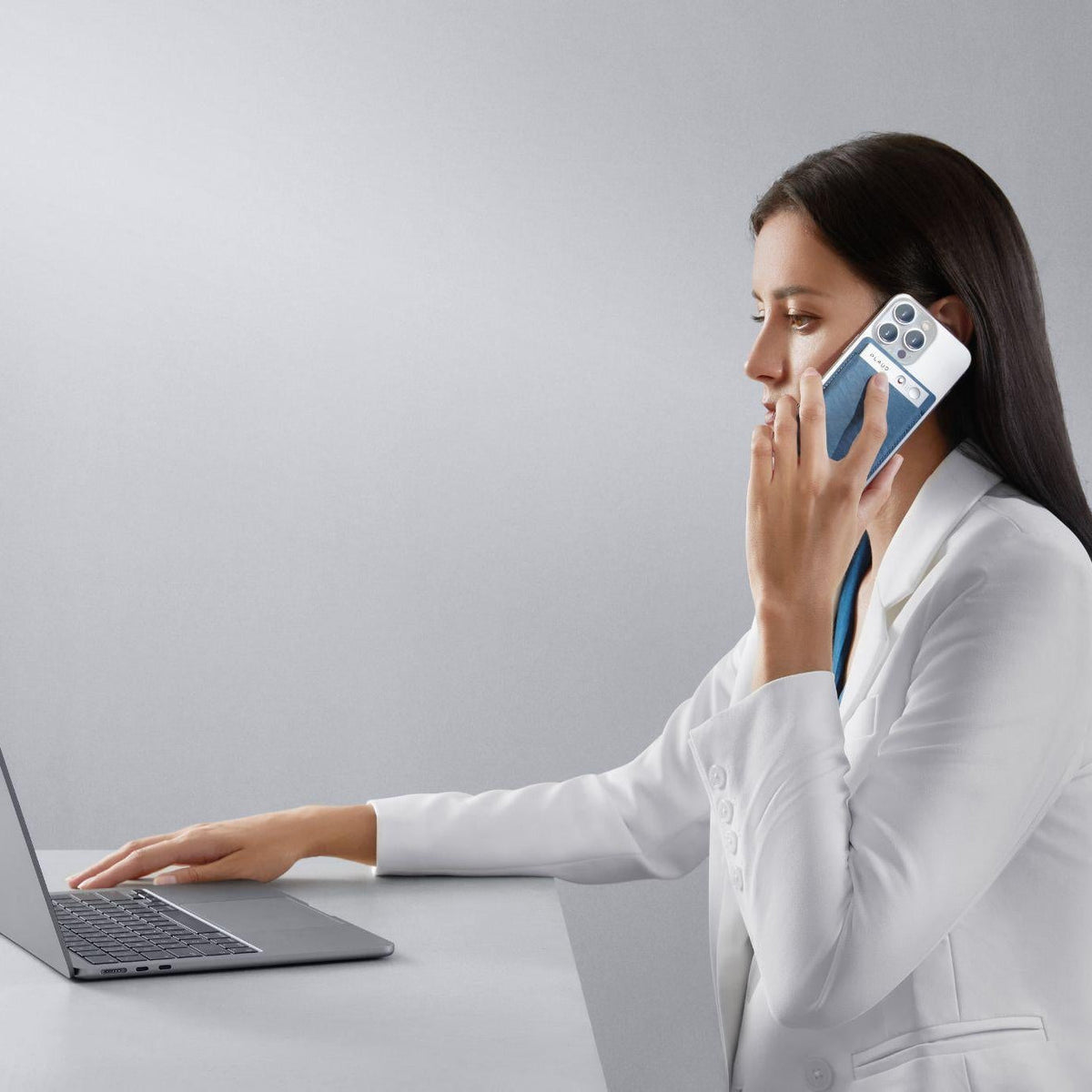 Woman using laptop while holding a phone with a blue Plaud Note attached to record a call.