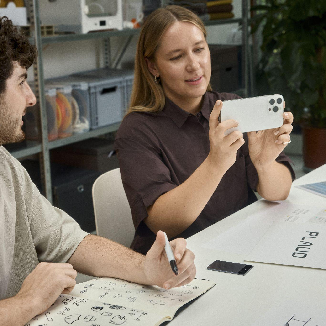 A black Plaud Note Pro on a table during a creative meeting while a woman takes a photo of sketches.