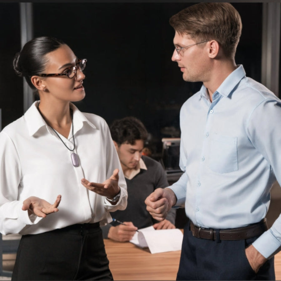 A woman wearing a purple Plaud NotePin on a lanyard talks to a man in an office setting, conveying professional discussion.