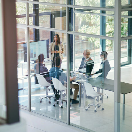 Executive team meeting in a glass-walled boardroom, with a presenter at a screen.