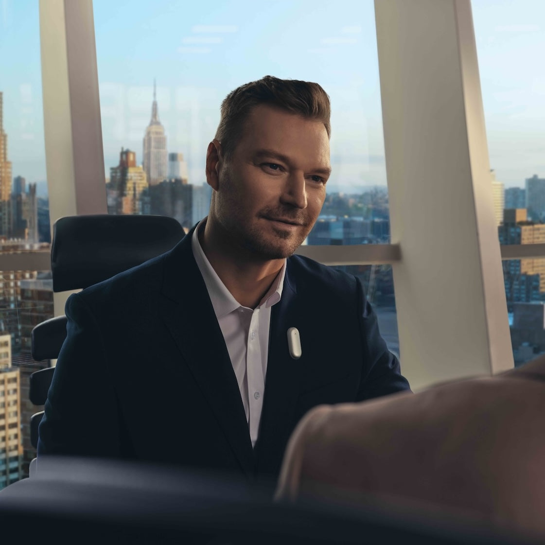 A man in a suit wearing a silver Plaud NotePin on his lapel during a meeting in a high-rise office.