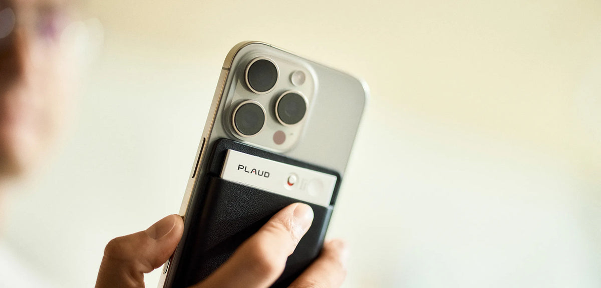 Black Plaud Note attached to a silver iPhone, with a finger pressing the device's button.