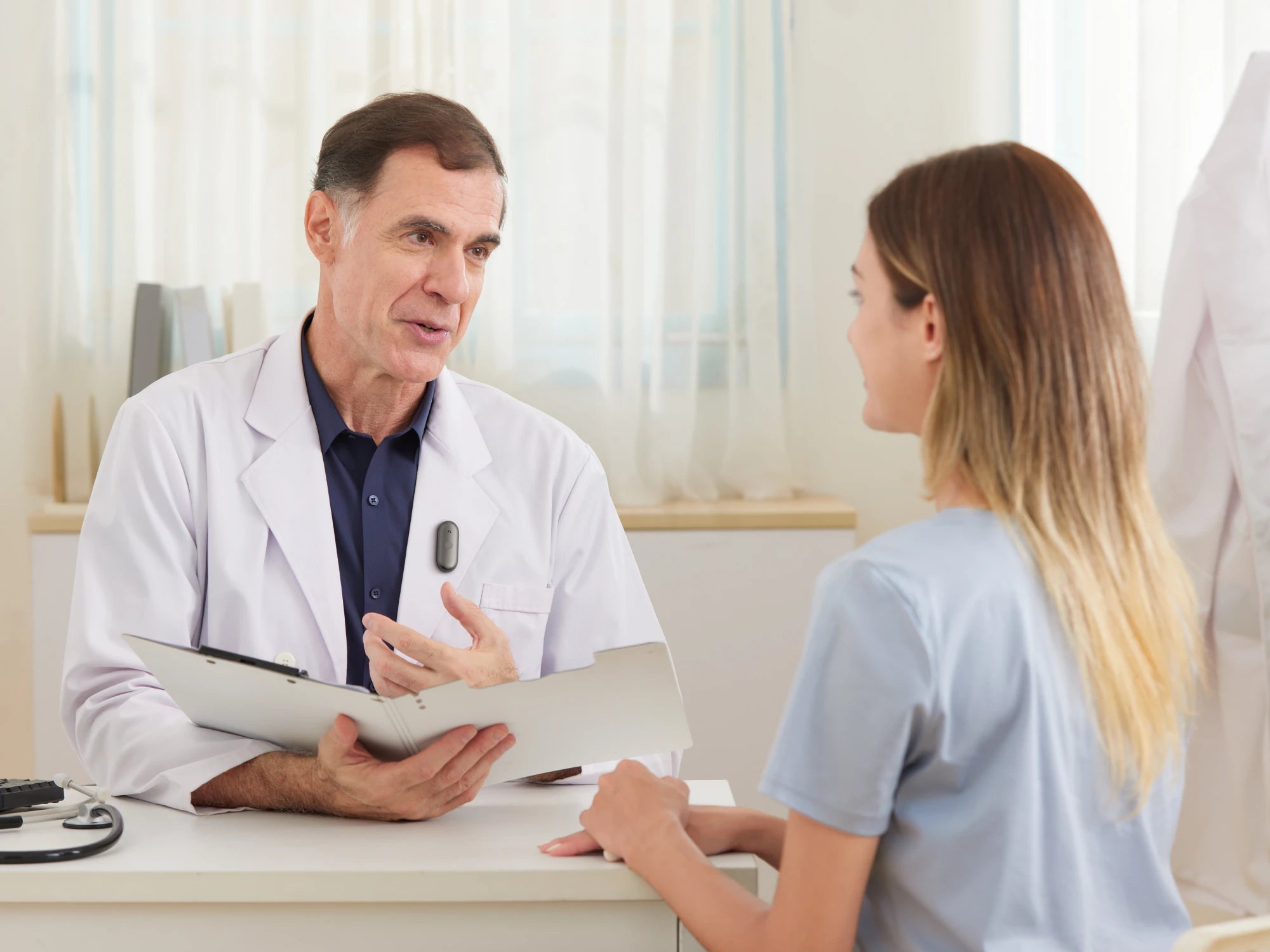 A doctor wearing a black Plaud NotePin S on his white coat while consulting with a patient.