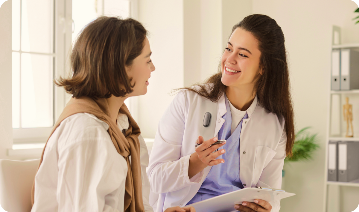 A smiling doctor wearing a Plaud NotePin S on her white coat speaks with a patient in a bright office.