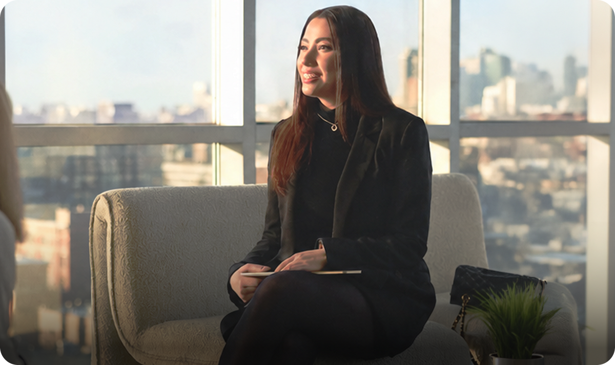 A professional woman in a black blazer sits on a couch in a high-rise office, holding a Plaud AI voice recorder during a meeting.