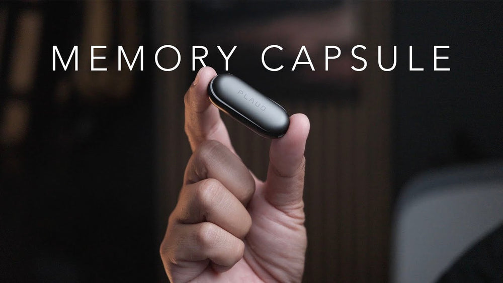 A hand holding a small, black Plaud NotePin wearable AI voice recorder with the text Memory Capsule.