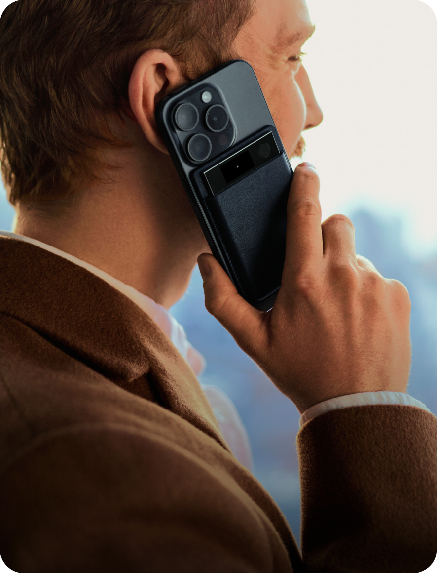 A man in a brown jacket holds a black smartphone to his ear with a black Plaud Note Pro attached to the back in a leather case.