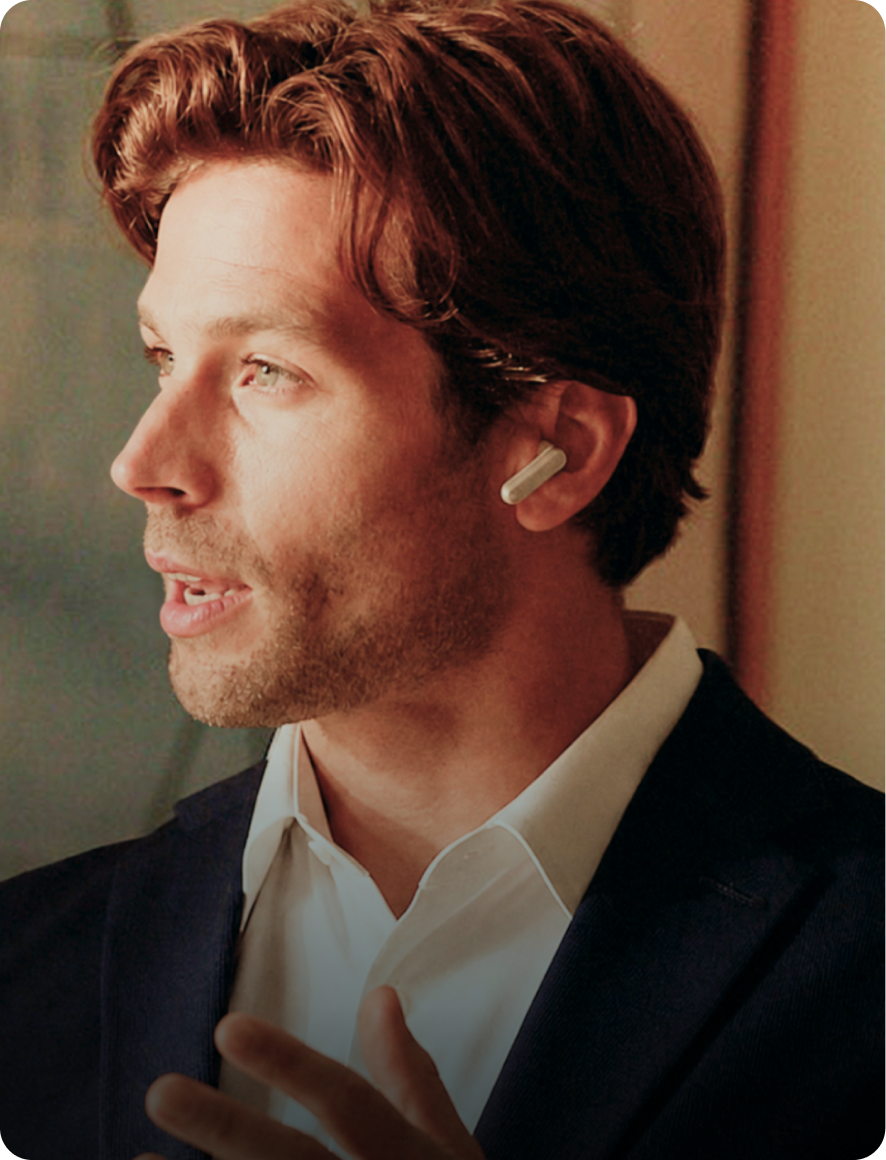 A man in a dark blazer and white shirt gestures while speaking, wearing a white Plaud One earbud with a silver stem.