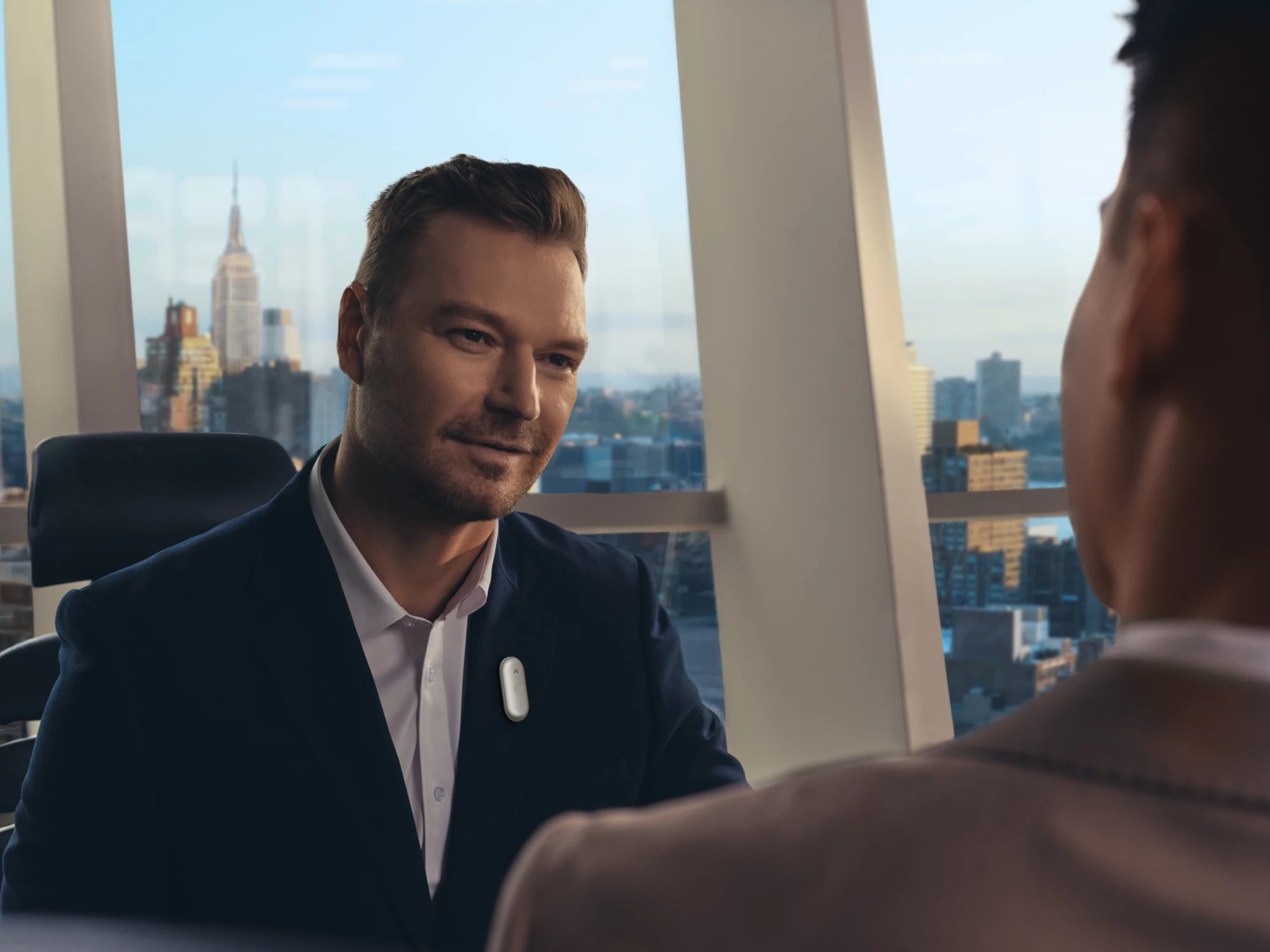 Professional man wearing a white Plaud NotePin S on his suit lapel during a meeting with a city skyline background.