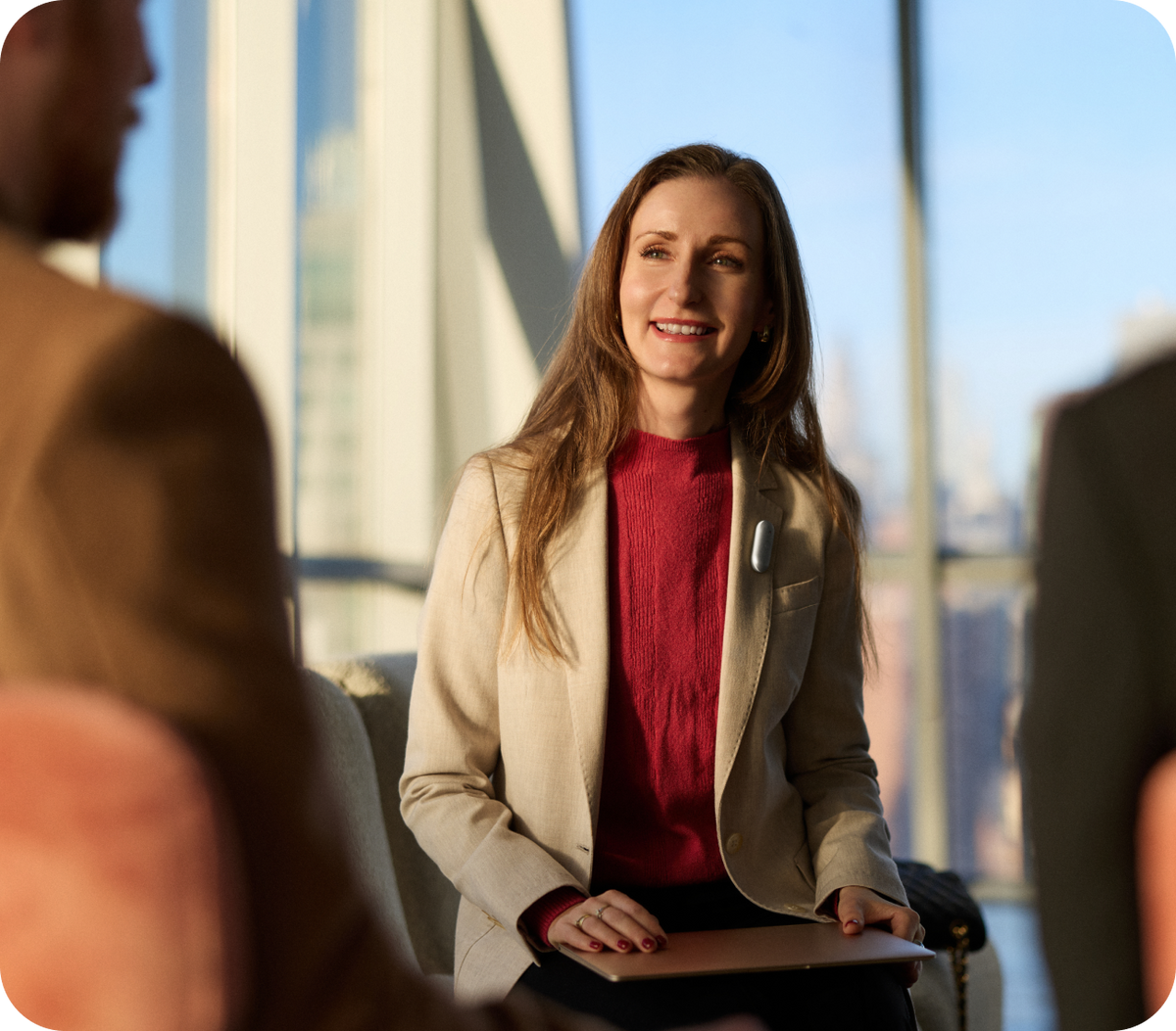 A smiling woman in a beige blazer and red turtleneck wears a silver Plaud wearable AI recorder during a meeting.
