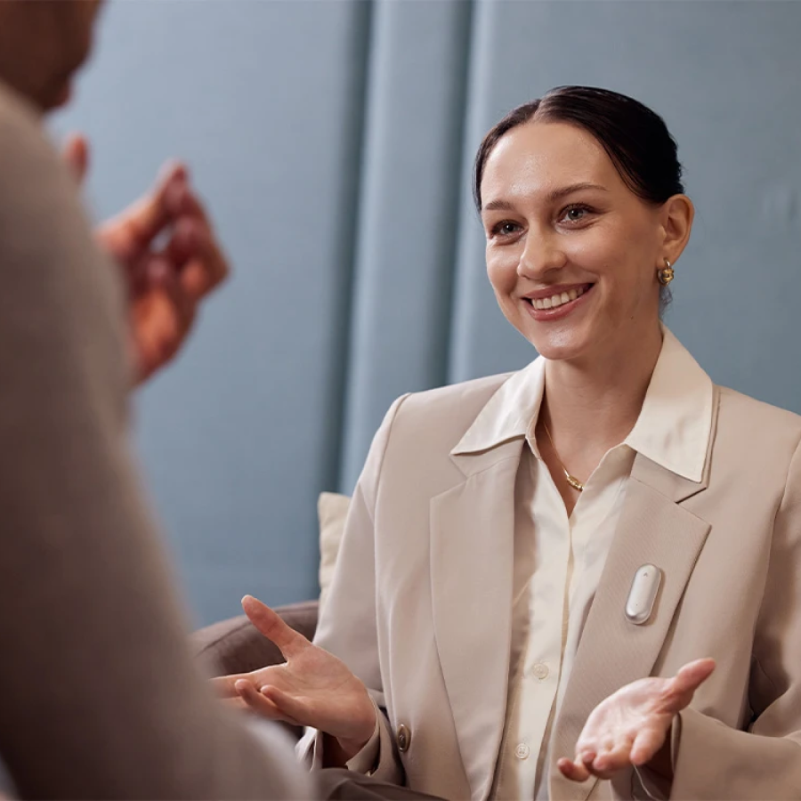 A woman wearing a silver Plaud NotePin S on her blazer lapel while smiling and talking during a meeting.
