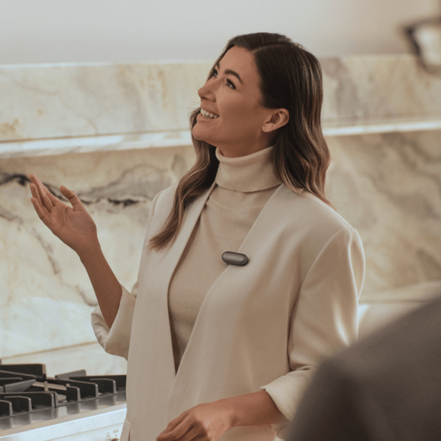 Woman wearing a gray Plaud NotePin on her lapel while speaking in a modern kitchen.