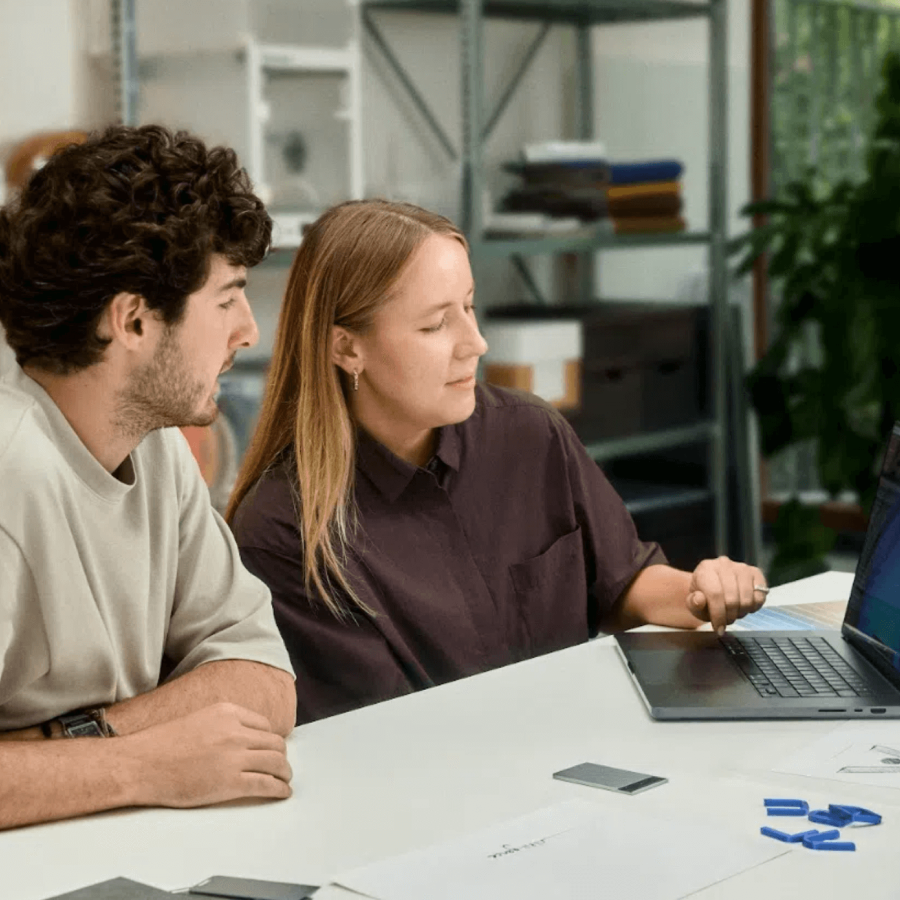 Two colleagues collaborating at a desk with a silver Plaud Note Pro AI voice recorder nearby.