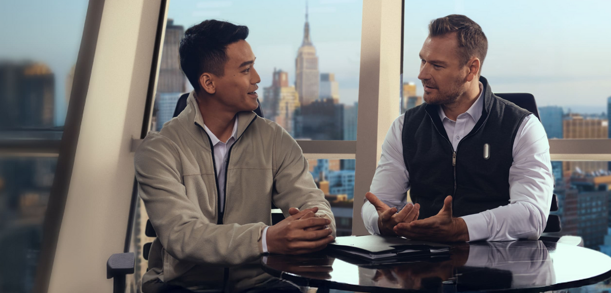 Two professionals in a meeting; the man on the right wears a silver Plaud NotePin on his black vest.