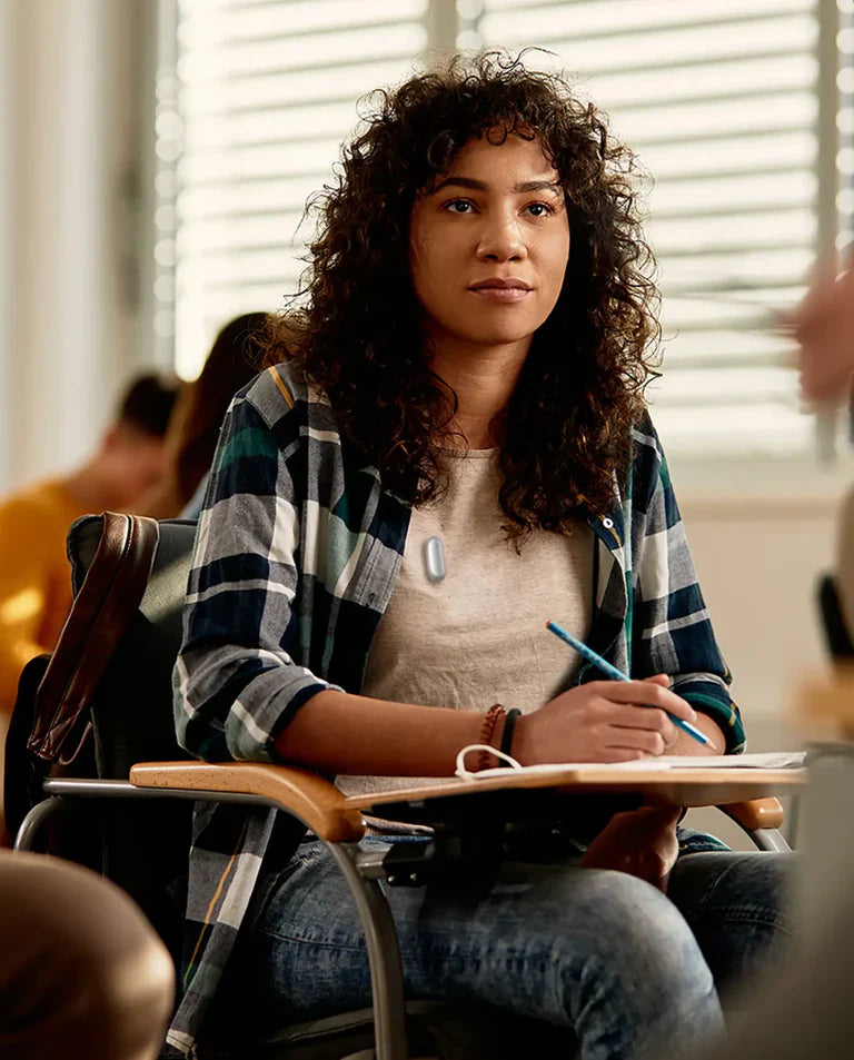 A woman in a classroom wearing a Plaud NotePin on her shirt while taking notes.