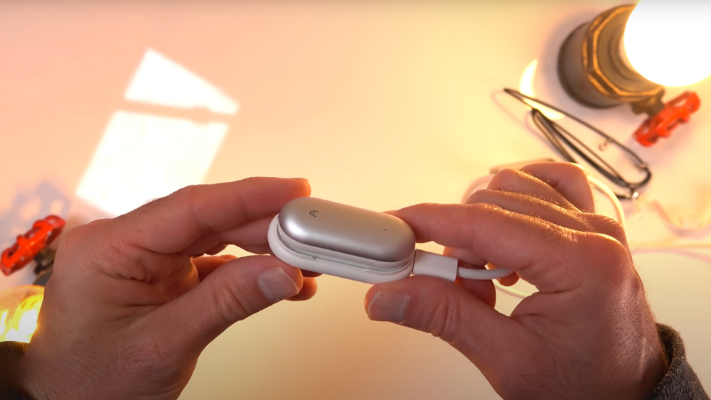 Hands holding a silver Plaud NotePin connected to a white charging cable under warm light.