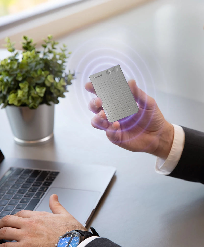 Hand holding a silver Plaud Note with purple signal waves over an office desk with a laptop.