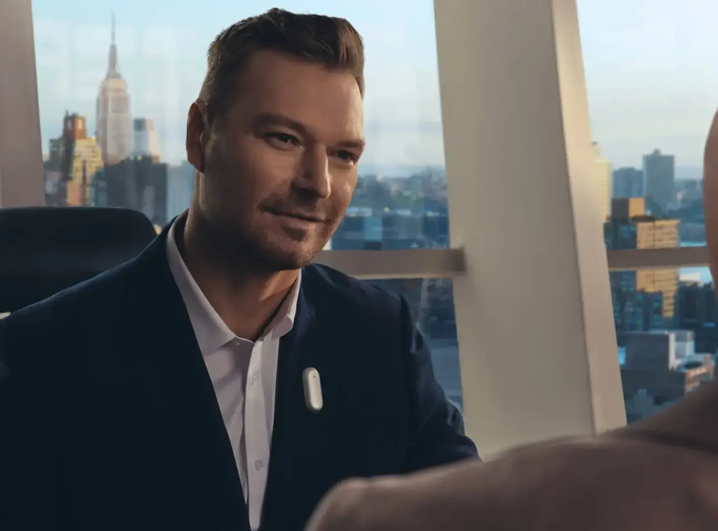 Man in a suit wearing a Plaud NotePin on his lapel during a meeting in a high-rise office.